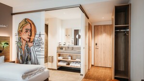 Una habitación de hotel moderna con una cama grande, un baño con tocador y estanterías, y un armario de madera.