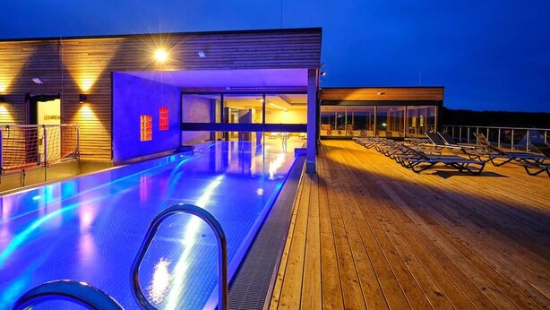 Zona de piscina moderna con terraza de madera y sillones de descanso, rodeada por un edificio con amplias ventanas y señalización iluminada.