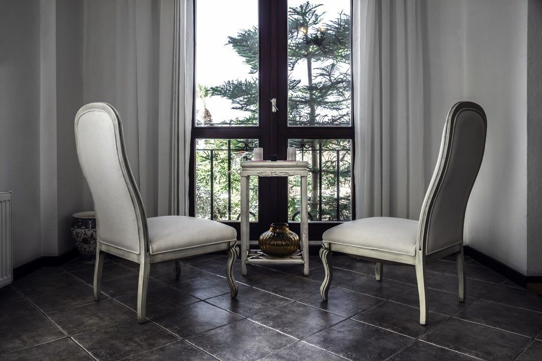 Two white upholstered chairs with curved legs, a small table with a vase, and a view of trees through a large window with curtains.