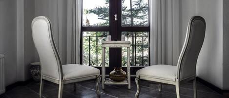Two white upholstered chairs with curved legs, a small table with a vase, and a view of trees through a large window with curtains.