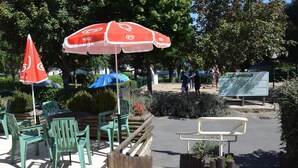 An outdoor seating area with green chairs and tables, red and white umbrellas, and a wooden bench. - Camping Parc de Charouzech (Salles-Curan)