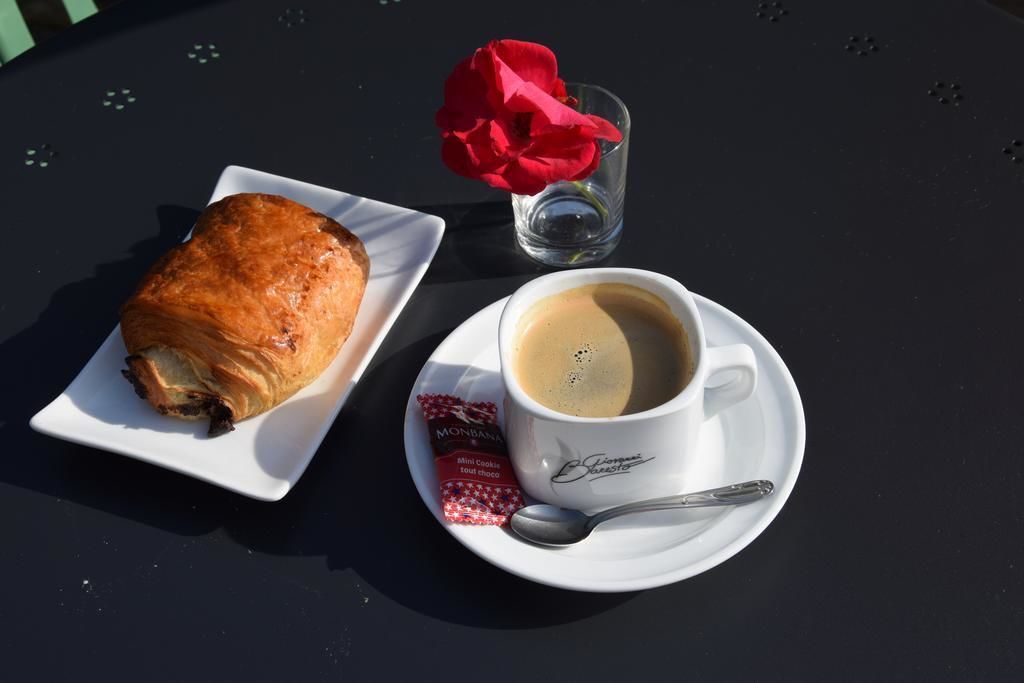 A croissant on a white plate, a cup of coffee with a spoon, and a glass with a red flower on a black table.