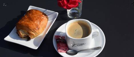 A croissant on a white plate, a cup of coffee with a spoon, and a glass with a red flower on a black table.