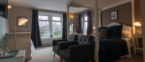 A four-poster bed with a dark blue bedspread, two armchairs, a bedside table with a lamp, and a window with curtains.