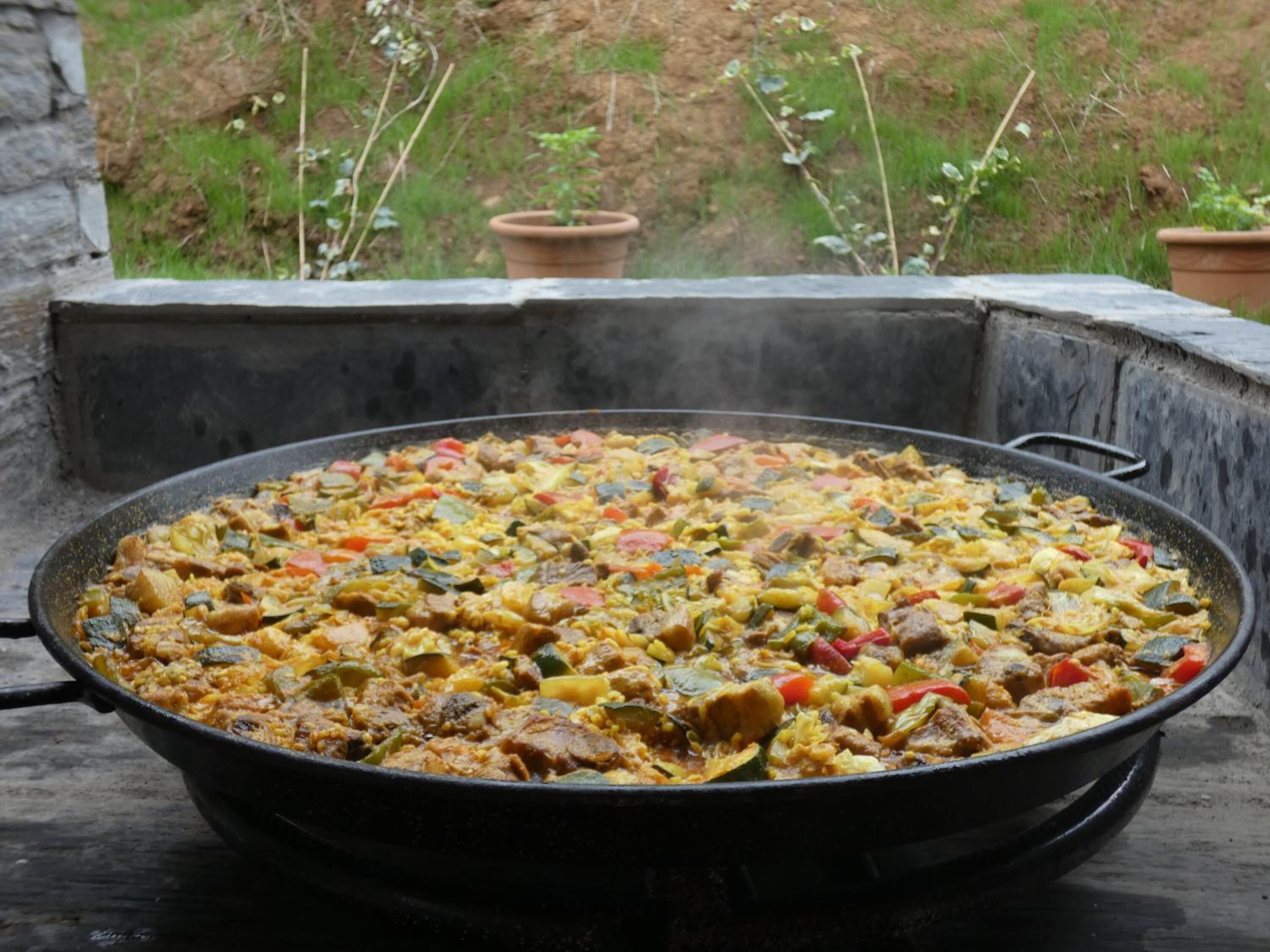 A large, round, black cooking pan filled with a colorful stew, placed outdoors on a stone surface with a garden view in the background.