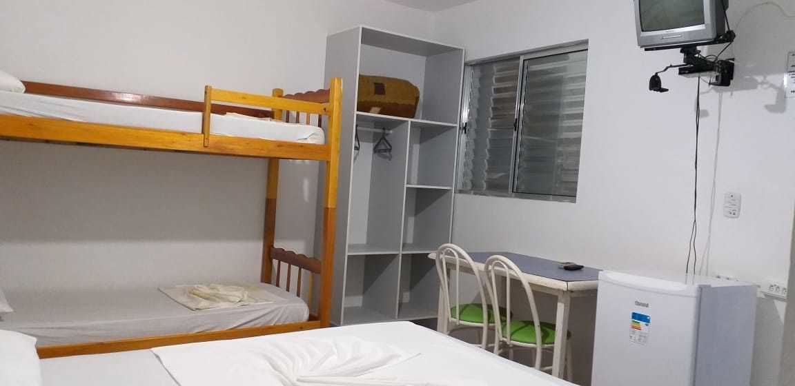A dormitory room with bunk beds, a desk, a chair, a small refrigerator, and a window with blinds.
