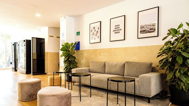 A modern lobby with a sofa, ottomans, and a coffee table. There are framed pictures on the wall and potted plants. - Micampus Málaga (Málaga)