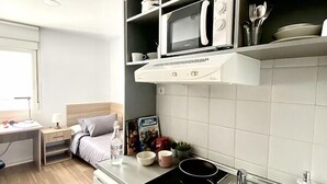 A compact kitchenette with a microwave, stove, and sink. There is a small dining area with a table and chairs, and a bed with a nightstand. - Micampus Málaga (Málaga)