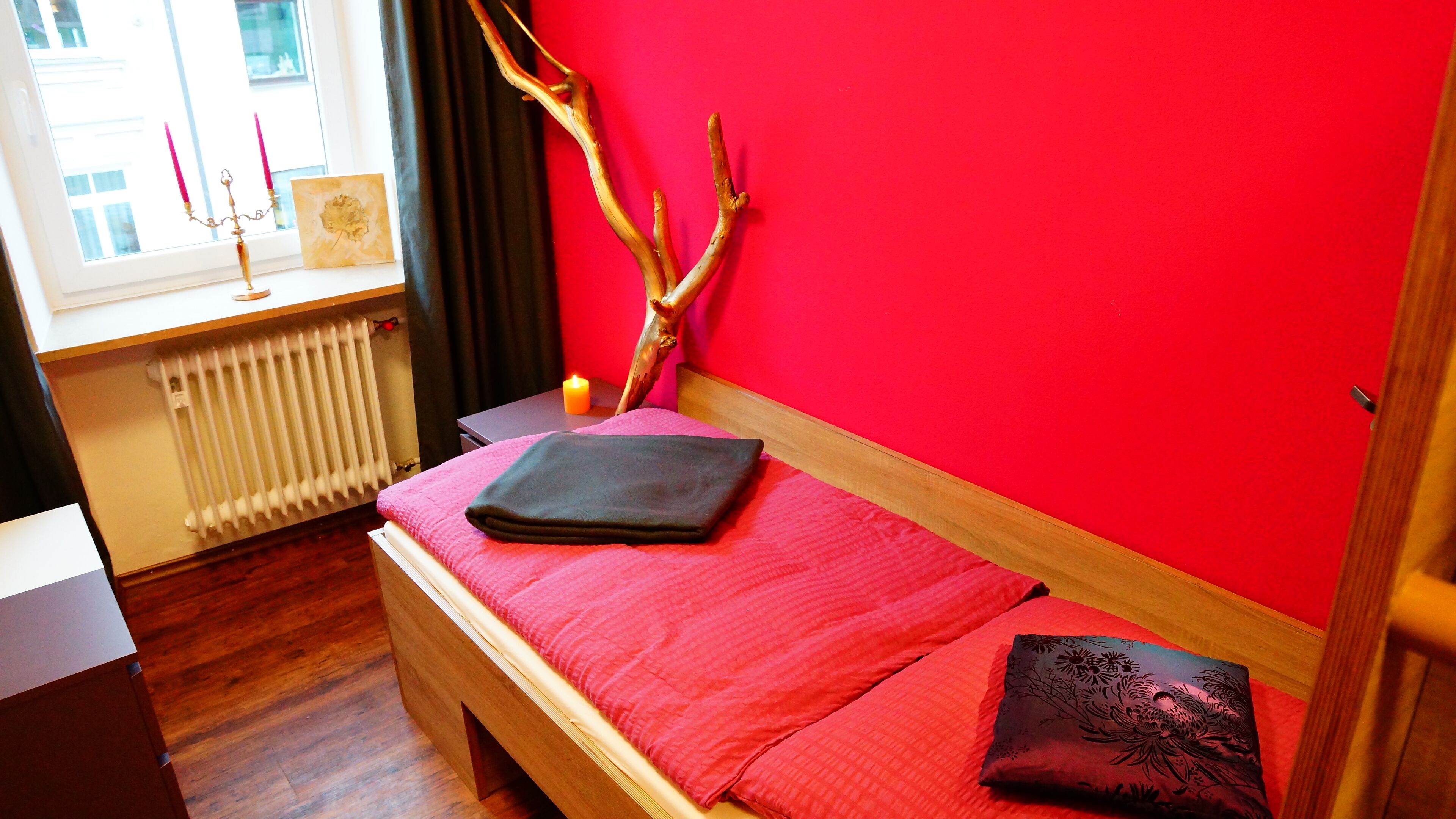 Ein Schlafzimmer mit einem hölzernen Bett, einer rosa Bettdecke, einem dunklen Kopfkissen, einer Zweigdekoration, einem Kandelaber mit Kerzen und einer Heizung.