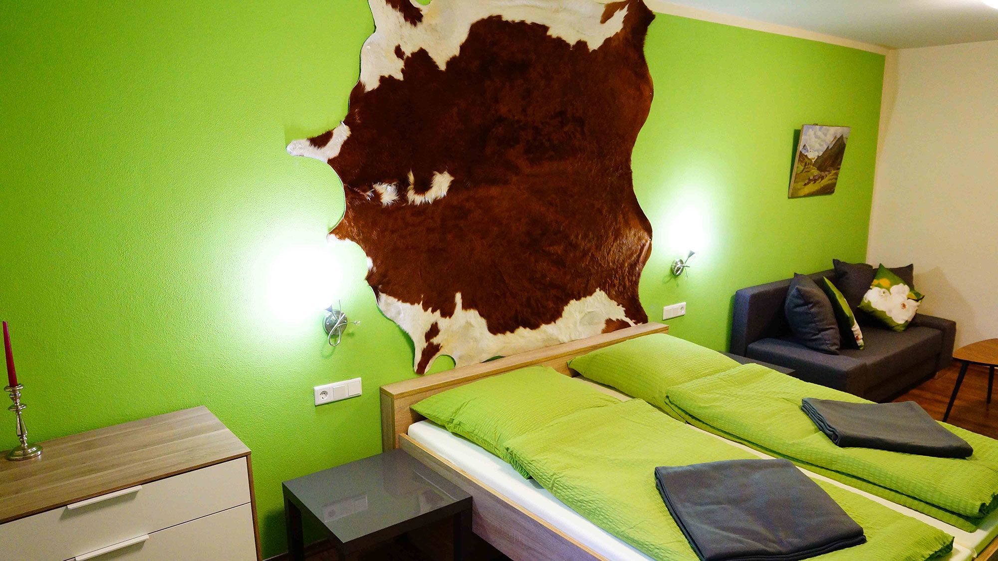 Ein Zimmer mit grüner Wand, einem großen Kunstwerk aus Kuhfell, einem Bett mit grüner Bettwäsche, einem kleinen Tisch, einem Nachttisch, einer Couch und einem Gemälde an der Wand.