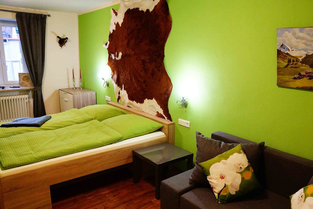 A room with a bed, a sofa, a green wall, a cowhide wall hanging, a radiator, and a painting of a mountain landscape.