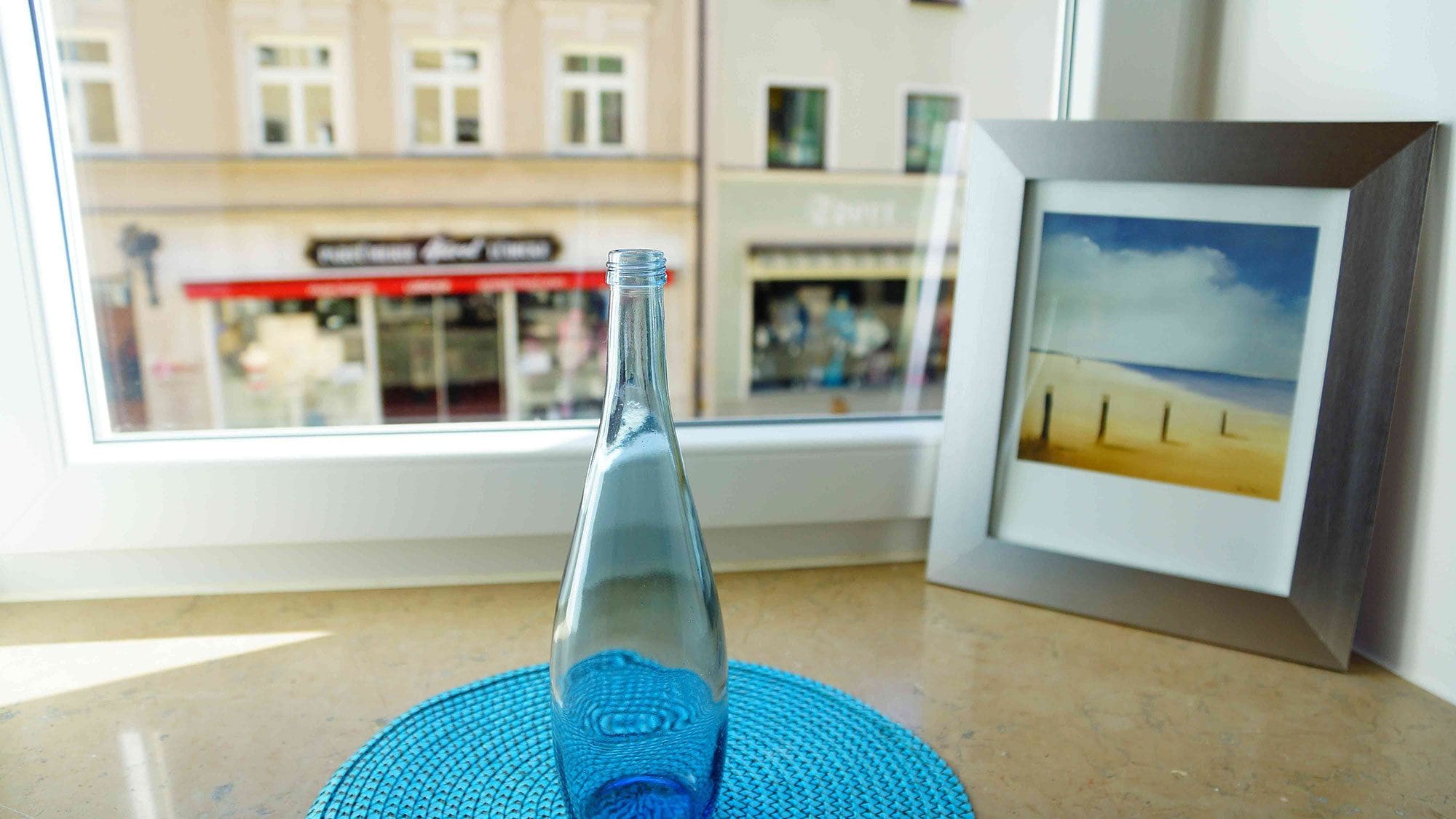A clear view of a bottle on a blue mat, a framed picture of a beach, and a window with a view of buildings and a store.