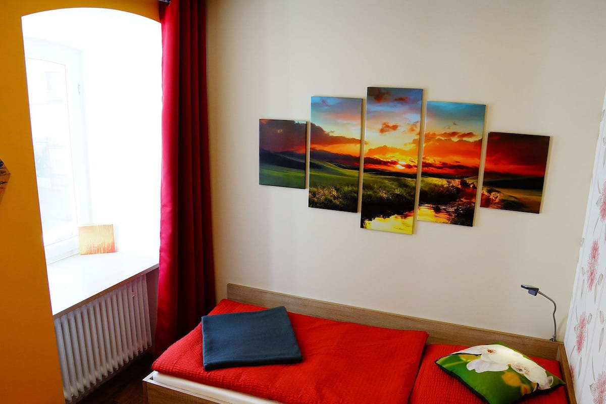 A room with a bed, a window with red curtains, a radiator, and a wall with a four-panel sunset painting.