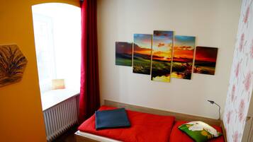 Ein Zimmer mit einem Bett, einem Fenster mit roten Vorhängen, einem Heizkörper und einer Wand mit einem vierteiligen Sonnenuntergangsgemälde.