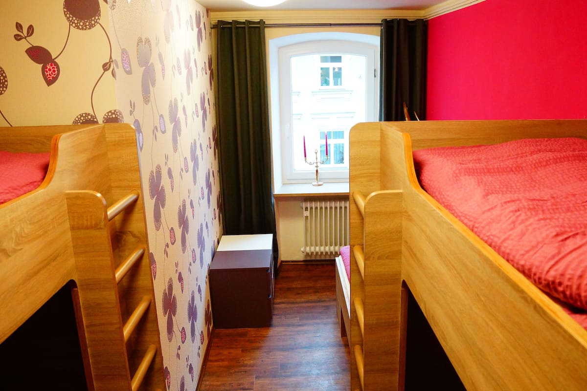 A room with two wooden bunk beds, a radiator, a window with curtains, and a wall with a floral pattern.