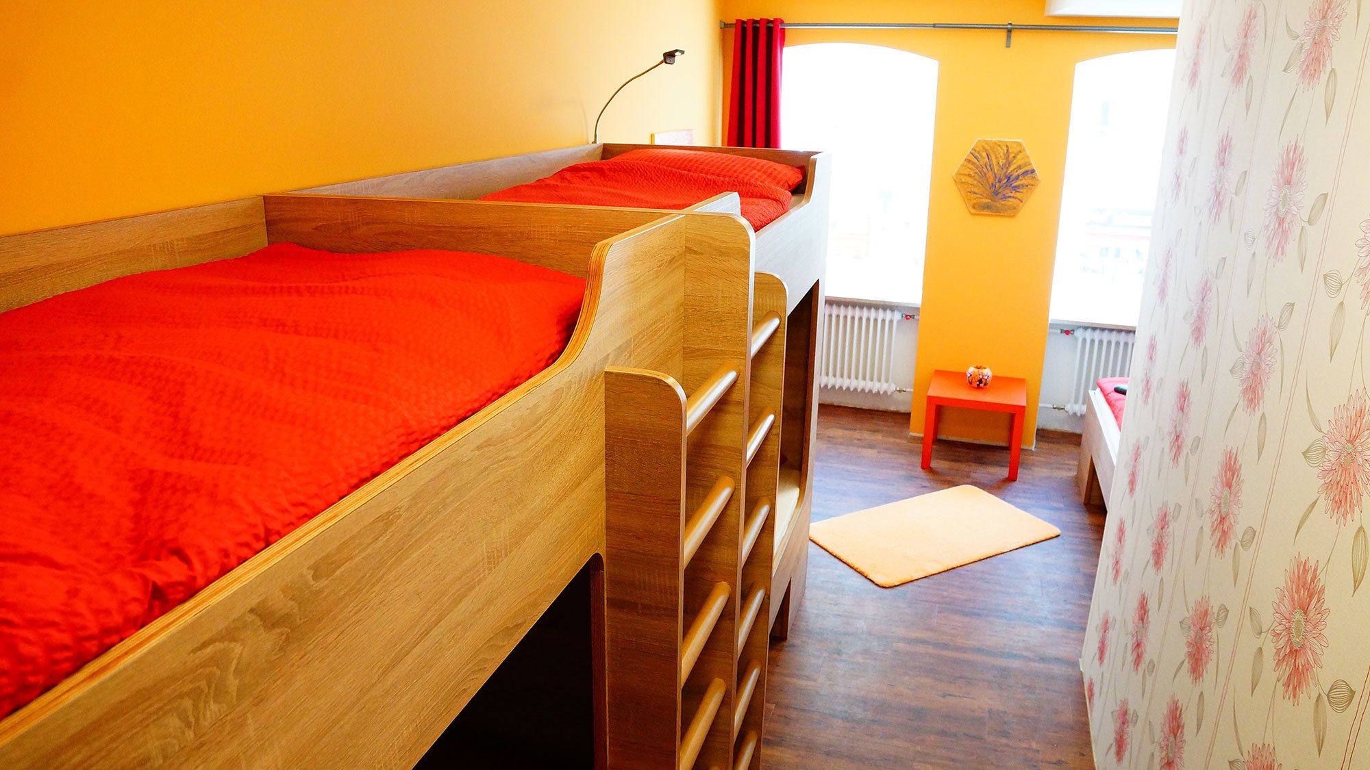 A room with two wooden bunk beds with red bedding, a small table with a red chair, and a window with red curtains.