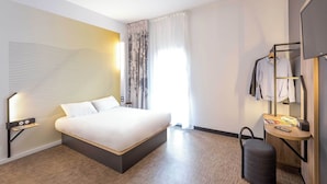 A modern hotel room with a large bed, a bedside table with a lamp, a small table with a mirror and a phone, a suitcase, and a wardrobe with hanging clothes. - B&B HOTEL SAINTE MAXIME Golfe de St Tropez (4628) (Sainte-Maxime)