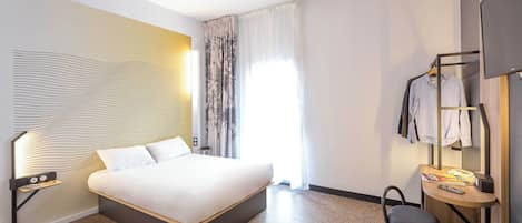 A modern hotel room with a large bed, a bedside table with a lamp, a small table with a mirror and a phone, a suitcase, and a wardrobe with hanging clothes.