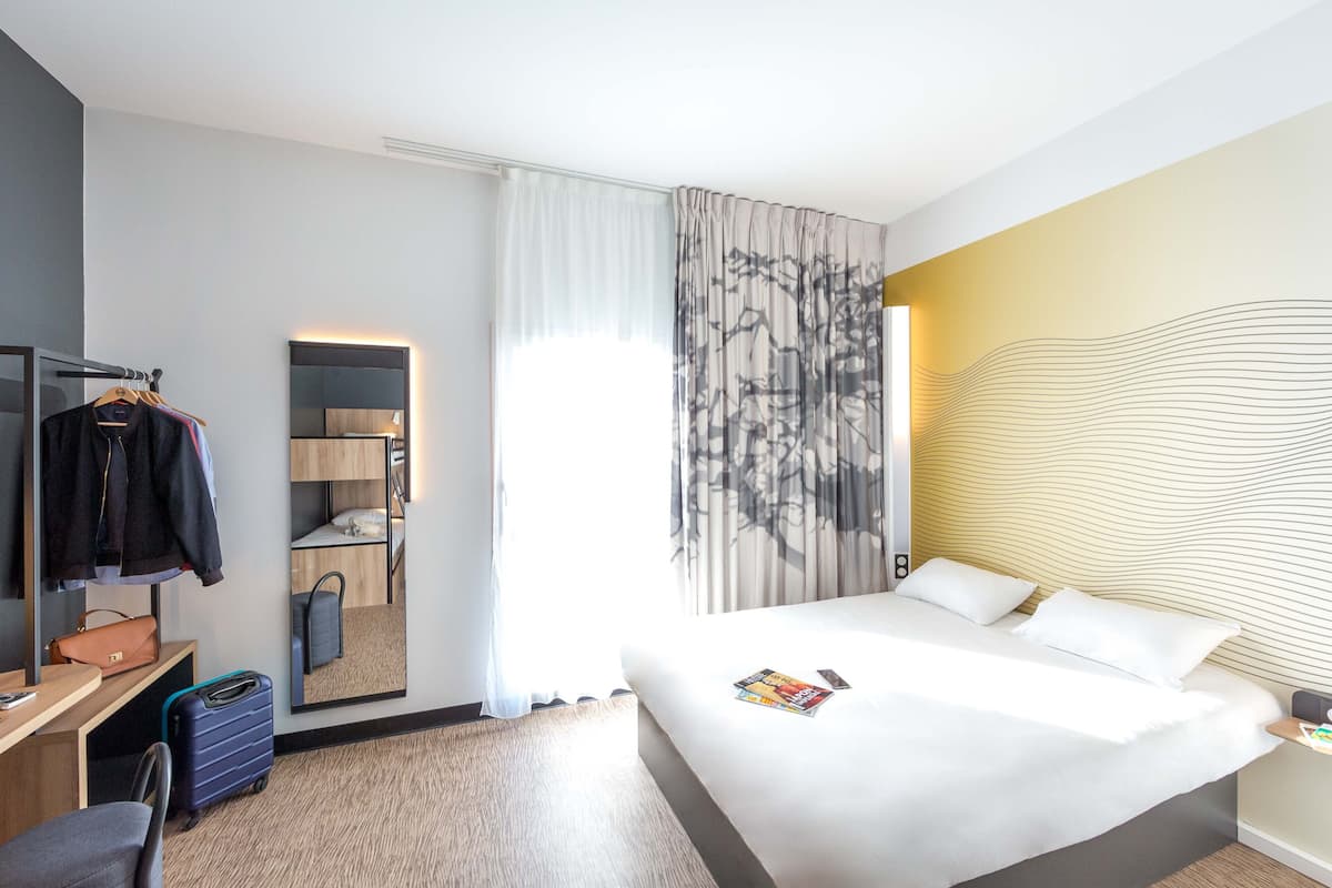 Ein Hotelzimmer mit einem Bett, einem Schreibtisch, einem Stuhl, einem Kleiderhaken, einem Koffer, einem Kleiderschrank und einem großen Fenster mit Vorhängen.