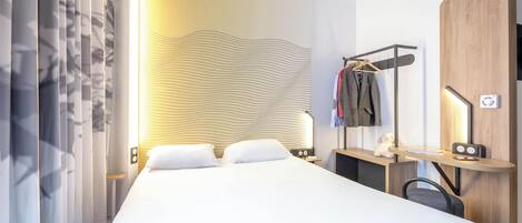 A hotel room with a bed, bedside table, lamp, and a coat rack with clothes.