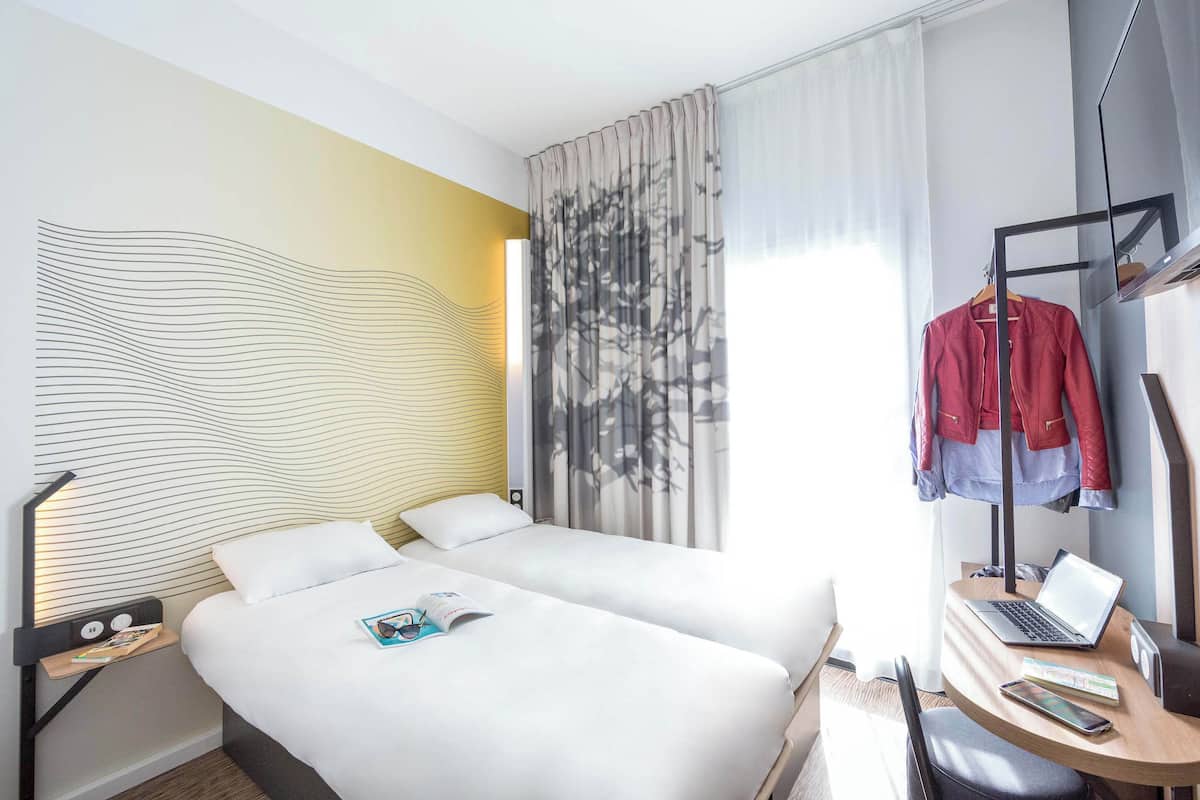 Ein Hotelzimmer mit zwei Betten, einem Schreibtisch mit Laptop, einem Kleiderständer mit Jacken und einem modernen Wanddesign.