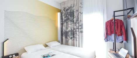 A hotel room with two beds, a desk with a laptop, a coat rack with jackets, and a modern wall design.