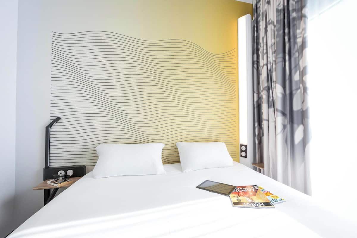Ein Hotelzimmer mit einem Bett, weißer Bettwäsche, einer Zeitschrift auf dem Bett, einem Nachttisch mit einer Lampe und einer Wand mit einem gewellten Muster.