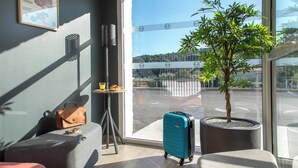 A modern hotel room with a large window, a potted tree, a blue suitcase, and a brown handbag on a sofa. - B&B HOTEL SAINTE MAXIME Golfe de St Tropez (4628) (Sainte-Maxime)