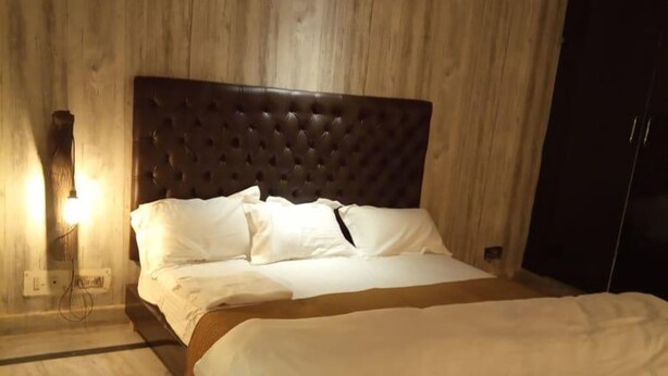 A bedroom with a wooden headboard, a bed with white linens, a bedside lamp, and a dark rug. - Rooming House (Greater Noida)
