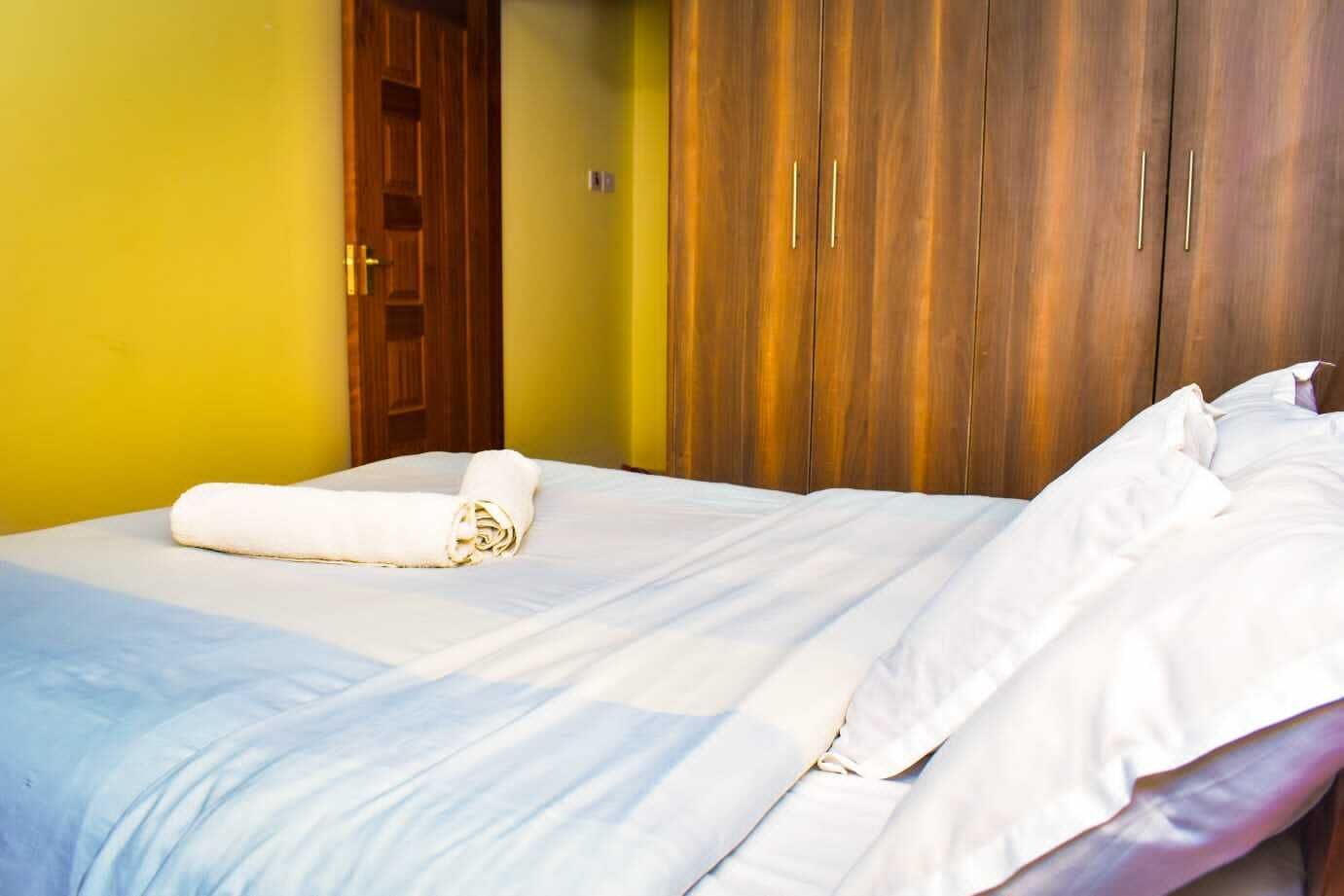 Ein ordentlich bezogenes Bett mit weißen Leinen und einem aufgerollten Handtuch, ein Holzschrank und eine Tür in einem Zimmer mit gelben Wänden.