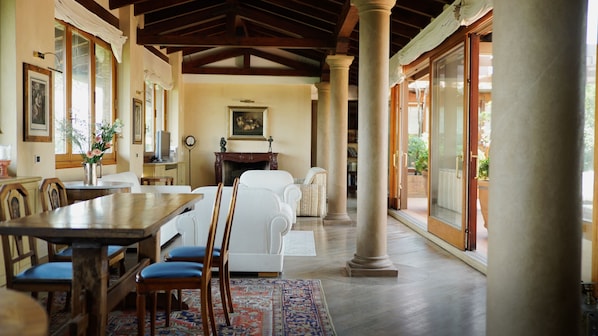 Cozy dining area with a fireplace, wooden table, and patterned rug, surrounded by framed artworks and a columned entryway.