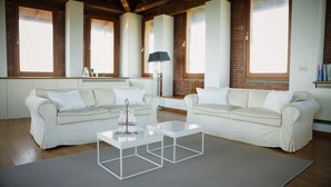 A spacious living room with two white sofas, a glass coffee table, and a lamp. Large windows provide natural light, and the room features exposed brick walls and wooden beams. - Antico Castello Marignolle (Florence)