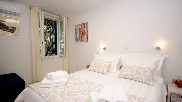 A neatly made bed with white linens and pillows, a window with curtains, a wall-mounted air conditioner, and a small table with a vase of flowers.