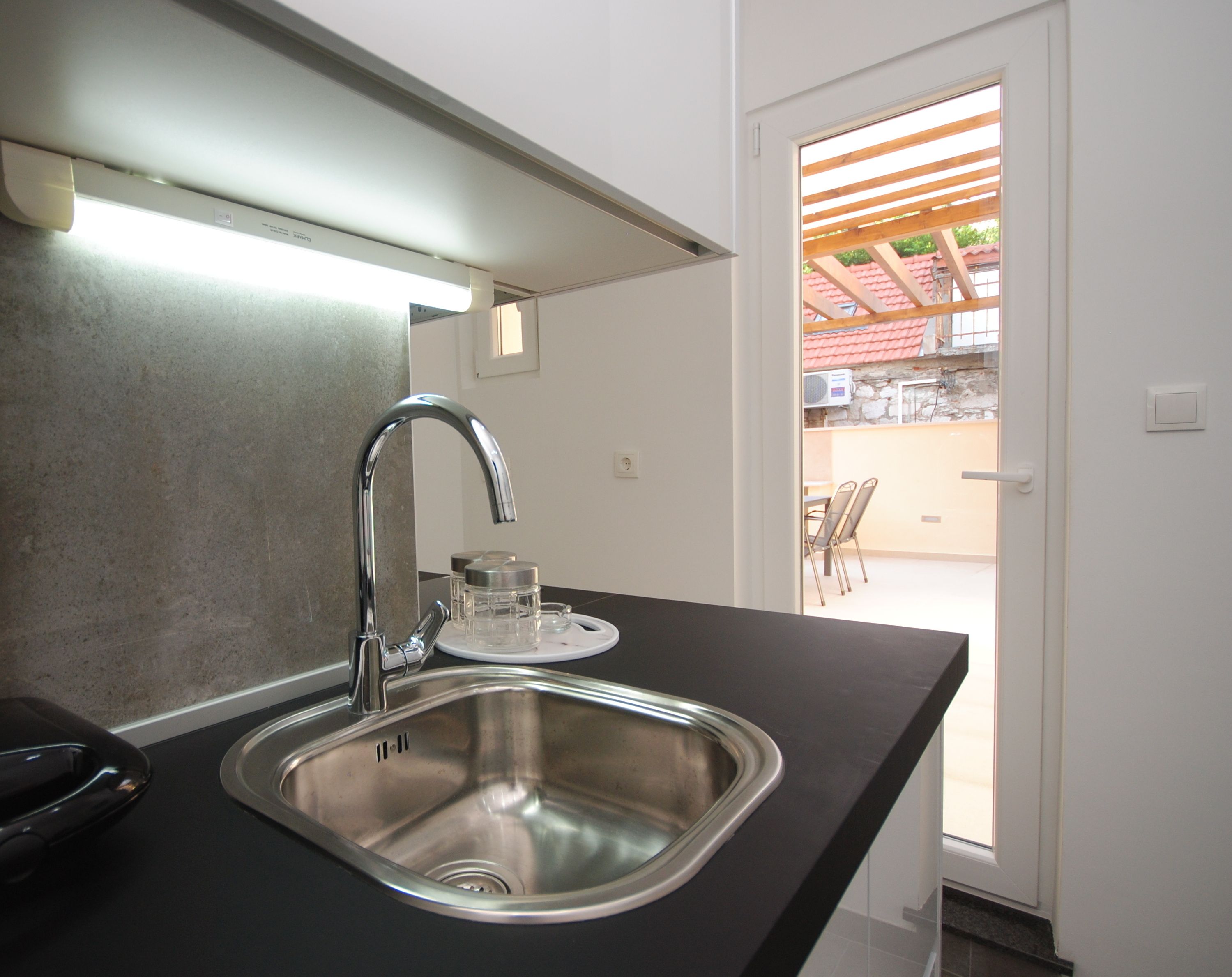 A modern kitchen with a stainless steel sink, a faucet, and a countertop. There is a window with a view of an outdoor patio area.