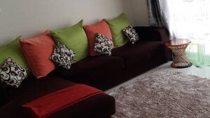 A living room with a dark sofa, green and red cushions, a patterned throw, and a small round table with a glass top. - Cafri Homestay (Syokimau)