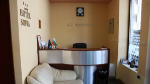 A reception area with a reception desk, a clock, a chair, and a trash bin. - Hotel Sofia (Pomorie)