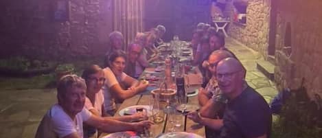 Un groupe de personnes est assis autour d’une table en bois à l’extérieur, la nuit, sous une guirlande lumineuse.