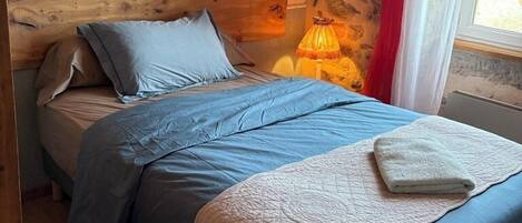 A bed with a blue and white quilt, a wooden headboard, a wall-mounted picture, and a stone wall with a red curtain.