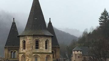 Un edificio histórico de piedra con una torre central y dos torres más pequeñas, rodeado de árboles y un paisaje montañoso brumoso.