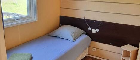 A compact bedroom with a single bed, a small bedside table, a window with curtains, and a built-in bench above the bed.