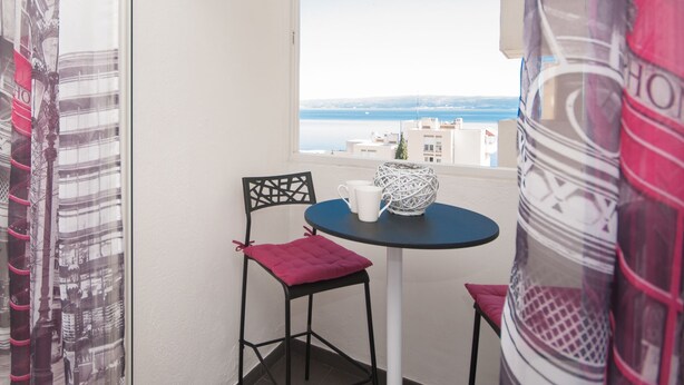 Ein kleiner Balkon mit Tisch und zwei Stühlen, Blick aufs Meer und ein Vorhang mit Muster.