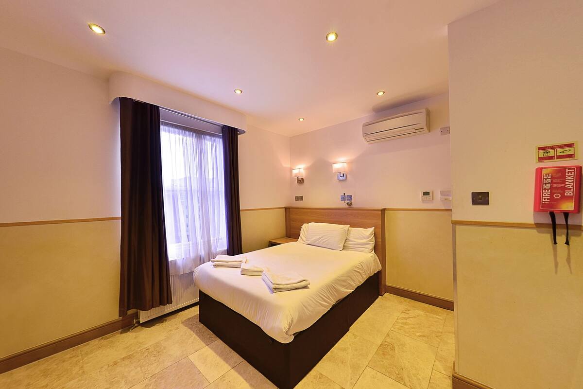 A hotel room with a bed, curtains, wall-mounted air conditioning, and a fire safety sign.