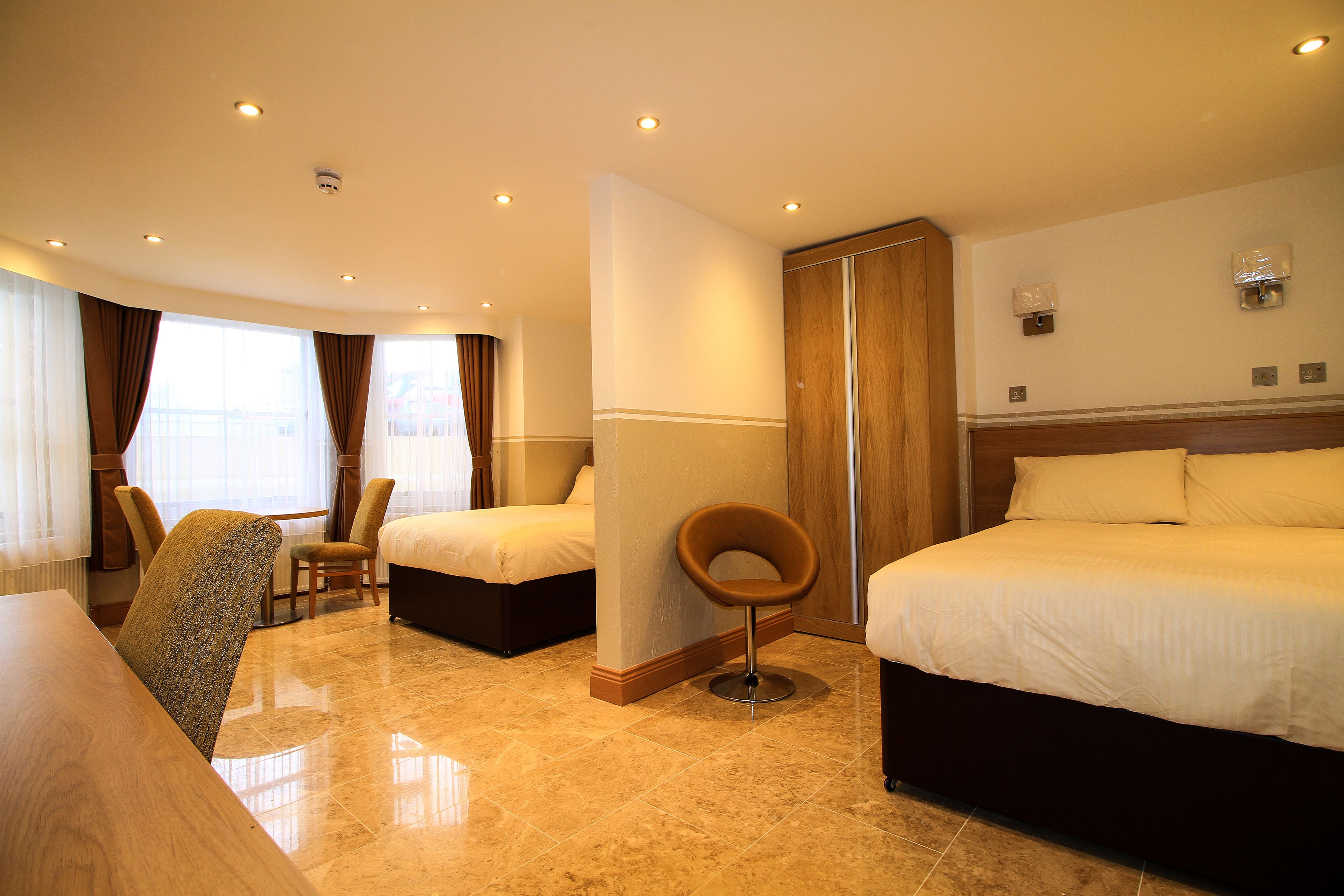 A hotel room with two beds, a sitting area with a chair, a wooden wardrobe, and a large window with curtains.