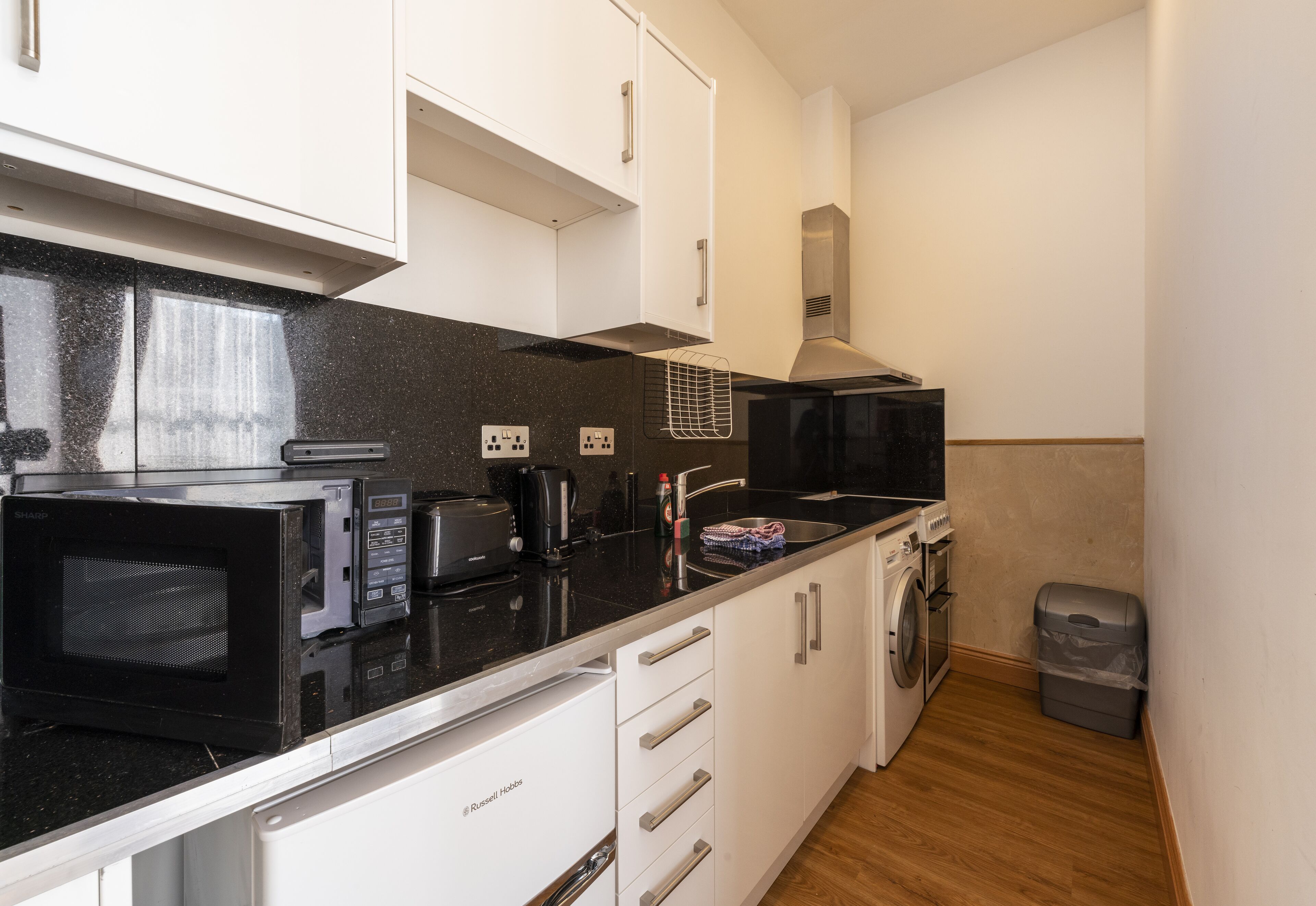 A compact kitchen with a microwave, toaster, and coffee maker on a black countertop. White cabinets and a stainless steel range hood are visible.