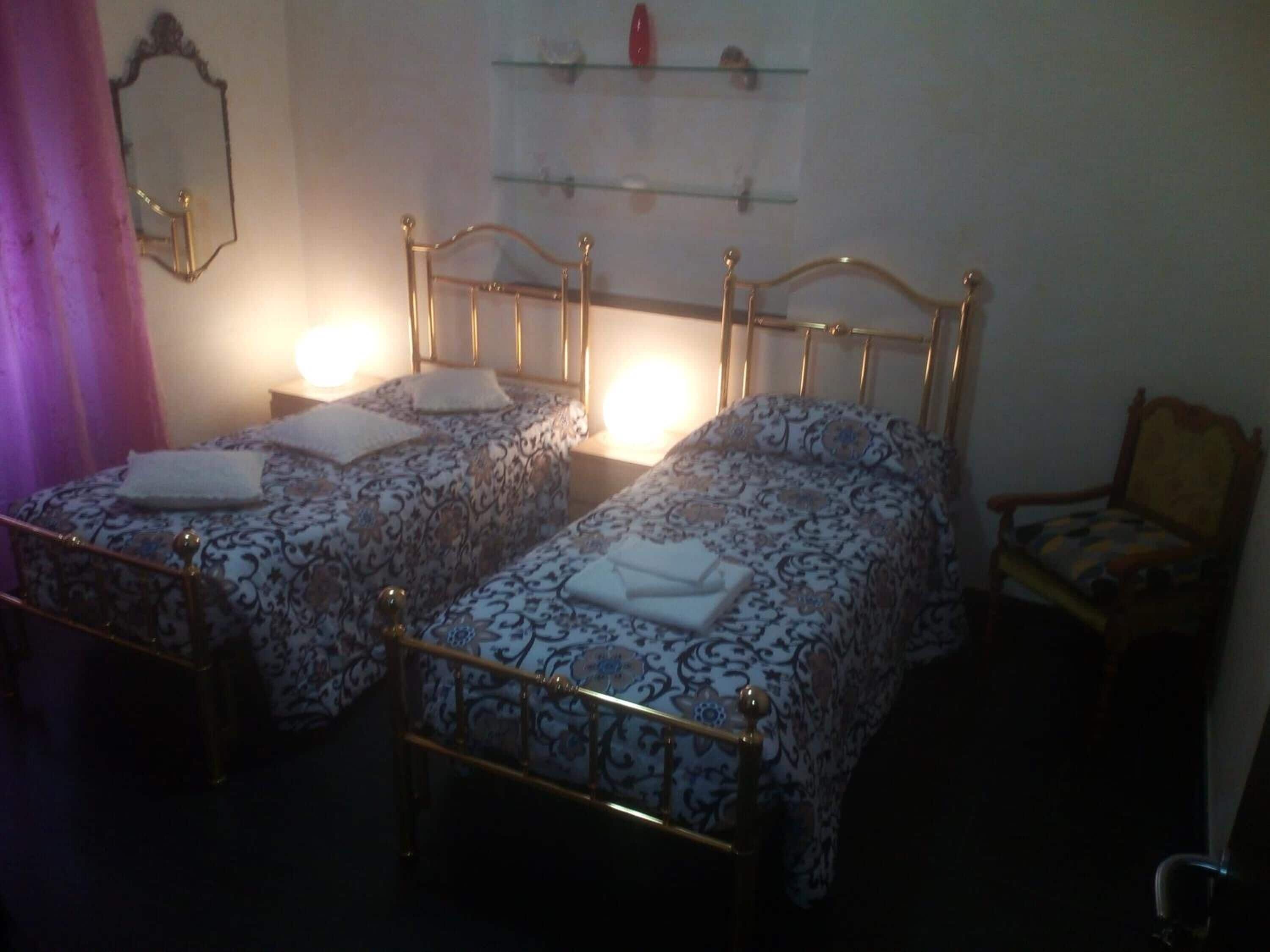 A room with two single beds with ornate metal frames, a wooden chair, and a wall-mounted shelf with decorative items.