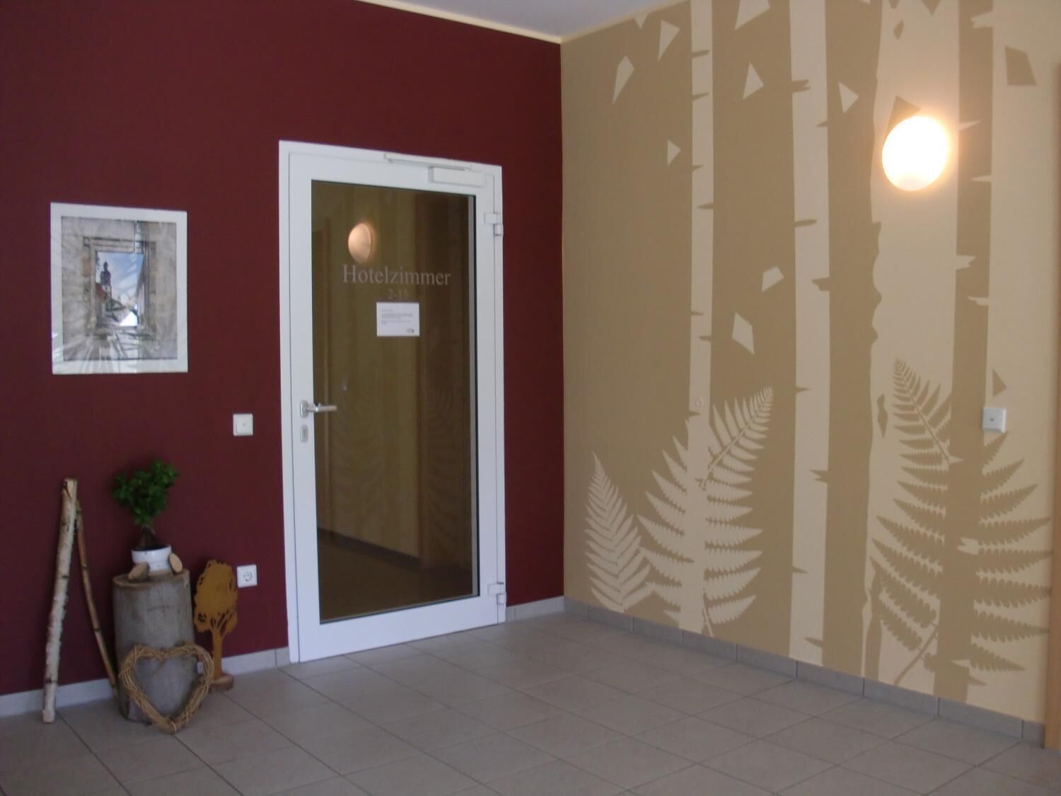 Habitación de hotel con una puerta que dice "Hotelzimmer 2-1-3", un cuadro en la pared, una planta pequeña y un árbol decorativo.