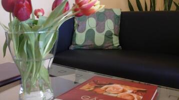 A clear glass vase with tulips on a table, a book titled 'GASTE und Freunde' in front of a sofa with a patterned cushion.