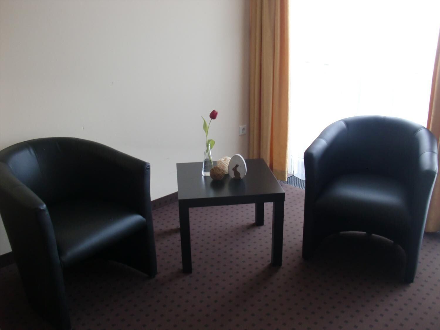 Una habitación con dos sillones de cuero negro, una mesita con un jarrón de una sola rosa roja y una ventana con cortinas.