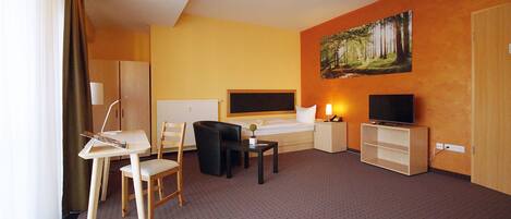 A hotel room with a bed, a desk with a chair, a TV, a painting on the wall, and a wardrobe.