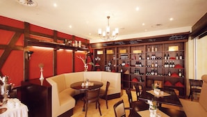 Un restaurante con una decoración en tonos rojo y marrón, que presenta una zona de bar con estantes llenos de botellas, un área de comedor con mesas y sillas, y un candelabro.